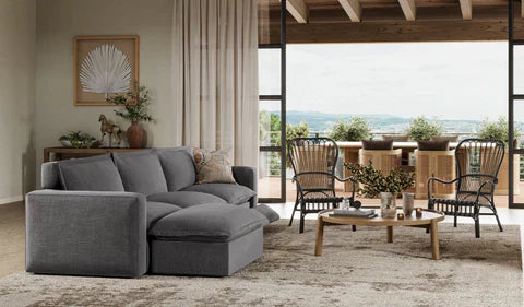 A cozy living room featuring a gray sectional sofa with a chaise, complemented by wooden furniture and large windows that offer a view of the outdoors.