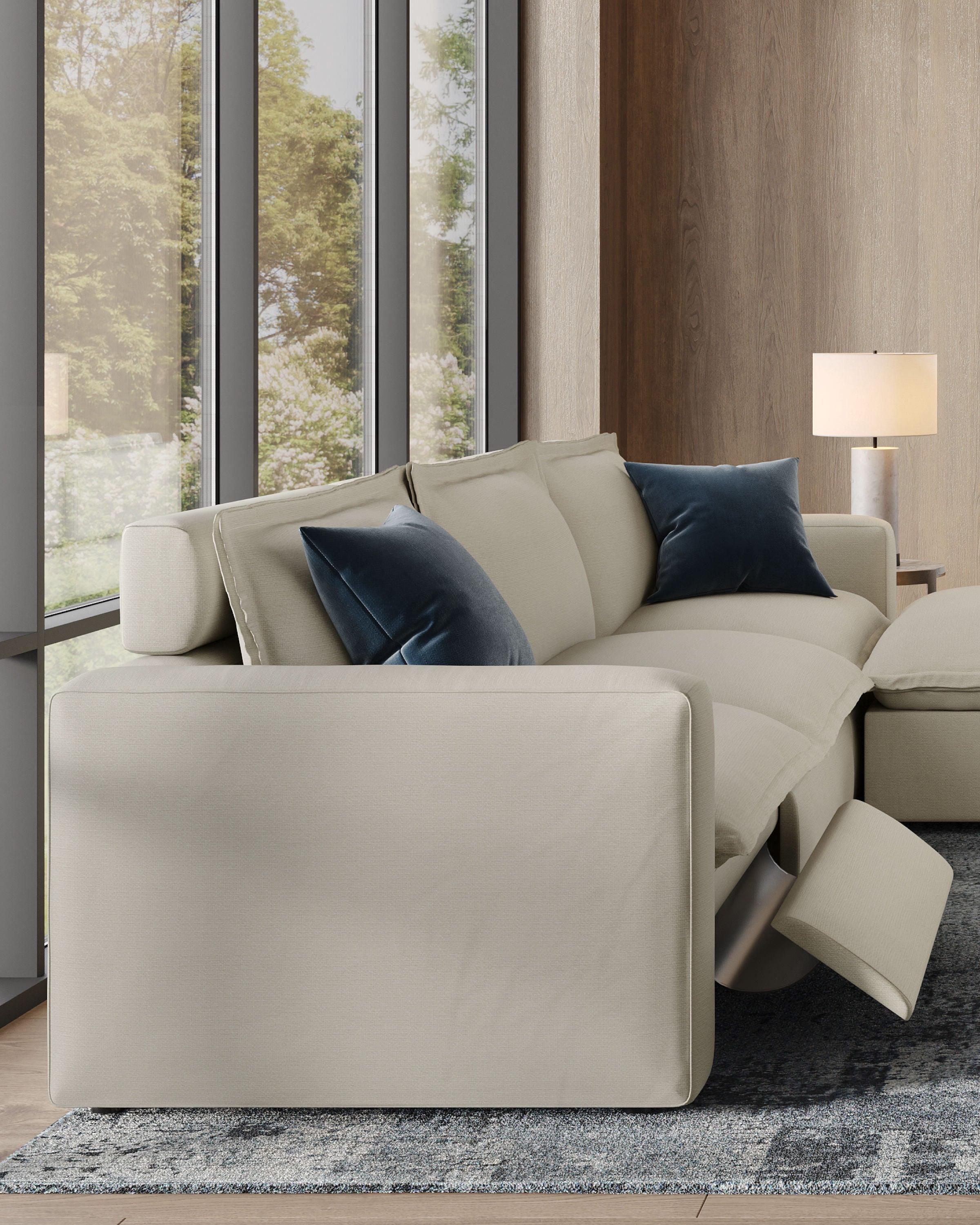 A close-up of a modern, light-colored reclining sofa with two dark blue pillows, set in a room with large windows. The leg rest of the sofa is partially extended