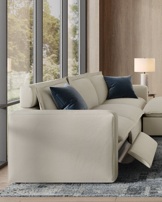 A close-up of a modern, light-colored reclining sofa with two dark blue pillows, set in a room with large windows. The leg rest of the sofa is partially extended