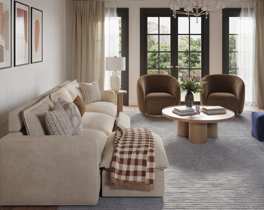 A stylish living room featuring a light-colored sectional sofa with decorative pillows and a plaid throw, two brown armchairs, and a round coffee table, all arranged on a blue-gray rug