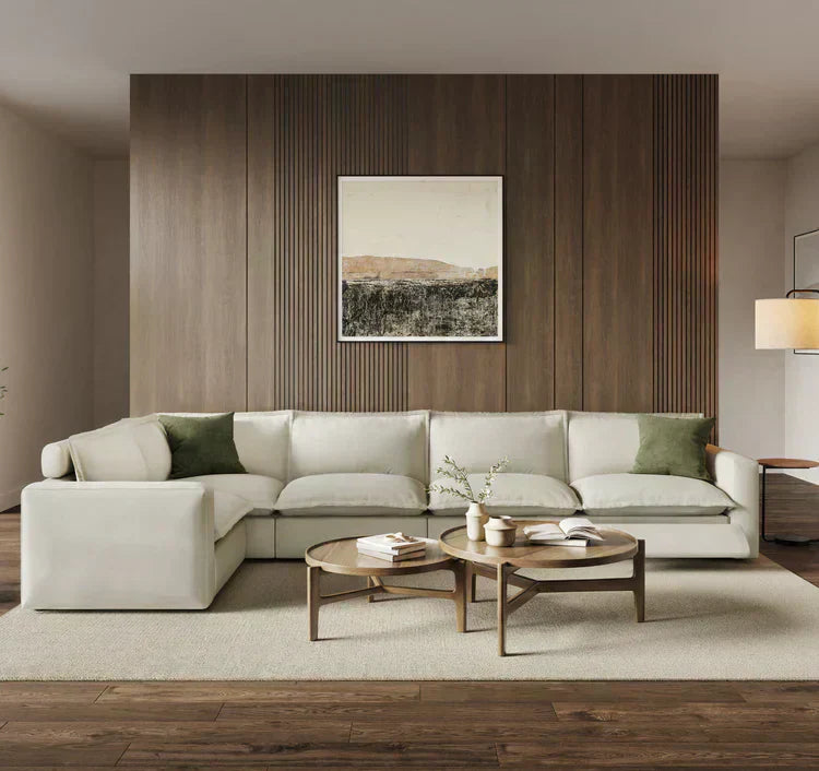 A modern living room featuring a large, off-white L-shaped sectional sofa with green accent pillows, set against a dark wood-paneled wall with framed art