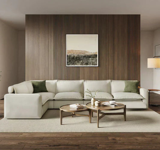 A modern living room featuring a large, off-white L-shaped sectional sofa with green accent pillows, set against a dark wood-paneled wall with framed art