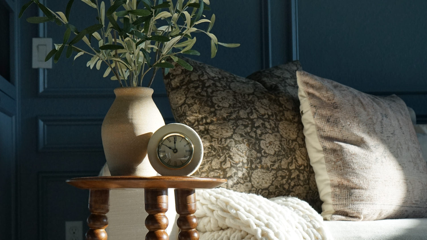 Cozy bedroom corner with deep blue paneled walls, featuring a small wooden side table with a ceramic vase, small clock, and decorative pillows with a white knit throw blanket