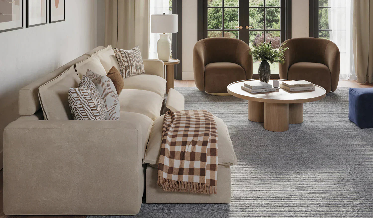 Cream-colored sectional sofa with decorative pillows in a contemporary living room featuring brown accent chairs, round wooden coffee table, and large windows with beige curtains