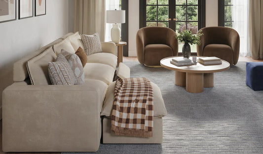 Cream-colored sectional sofa with decorative pillows in a contemporary living room featuring brown accent chairs, round wooden coffee table, and large windows with beige curtains